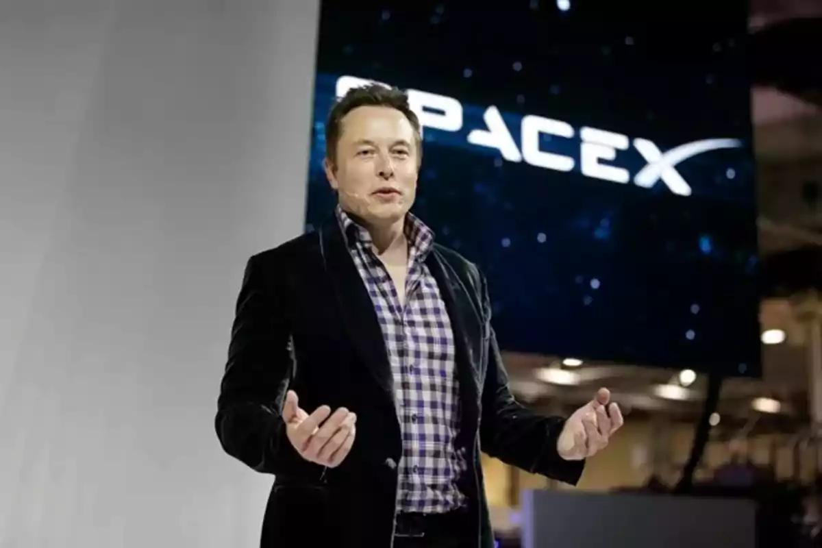 A man standing in front of a SpaceX sign, wearing a dark jacket and a checkered jersey.