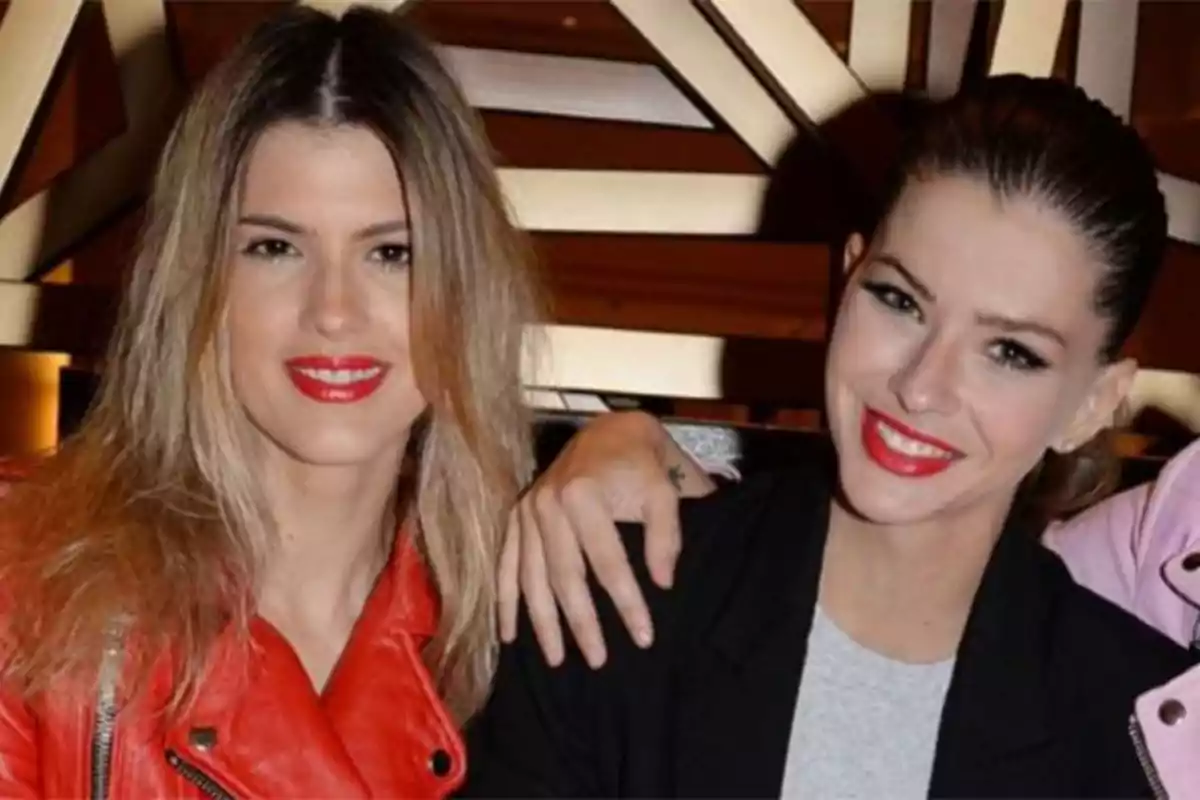 Dos mujeres jóvenes sonríen a la cámara, una lleva chaqueta roja y la otra chaqueta negra, ambas con labios rojos y fondo geométrico. Dos mujeres jóvenes sonríen a la cámara, una lleva chaqueta roja y la otra chaqueta negra, ambas con labios rojos y fondo geométrico.