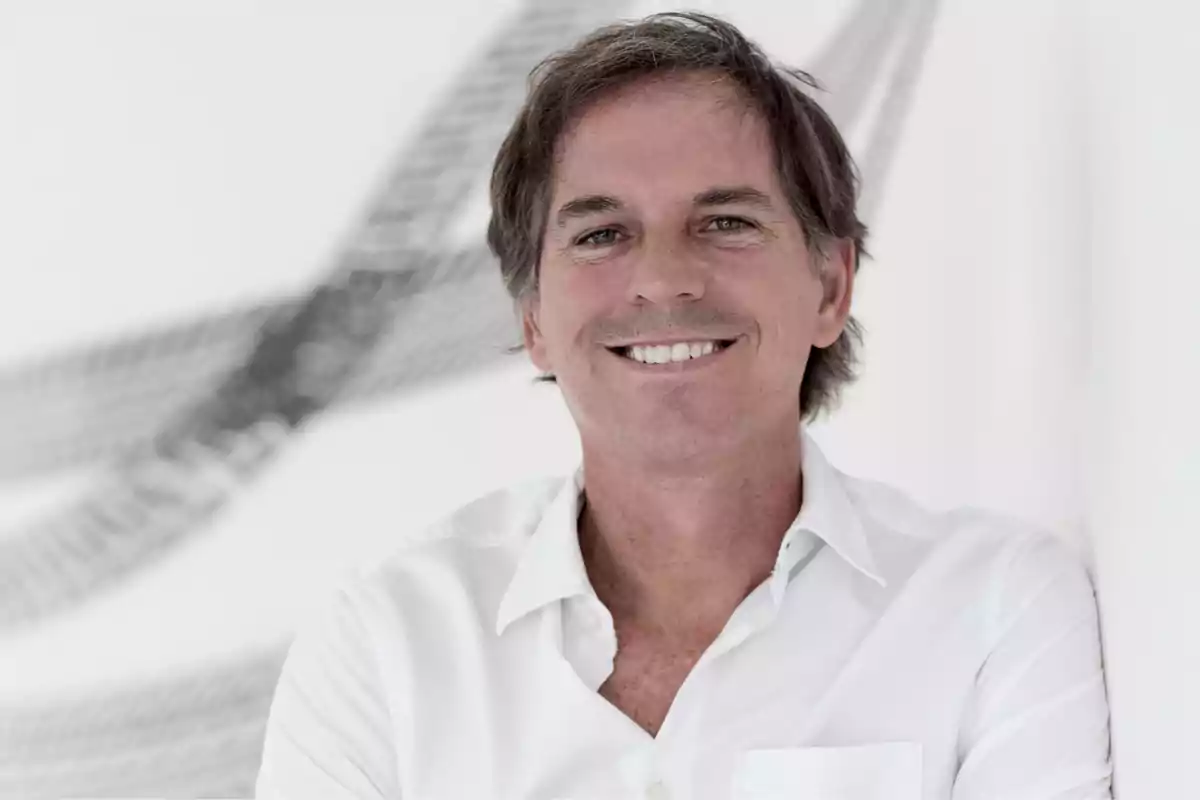 Hombre de cabello castaño y camisa blanca sonriendo frente a una pared blanca con un diseño abstracto en el fondo Hombre de cabello castaño y camisa blanca sonriendo frente a una pared blanca con un diseño abstracto en el fondo