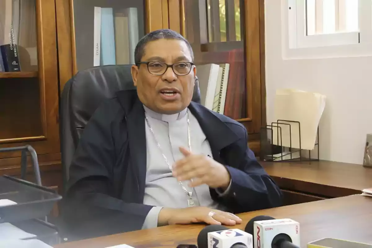 Un hombre con gafas y vestimenta religiosa está sentado en una oficina hablando frente a varios micrófonos sobre un escritorio