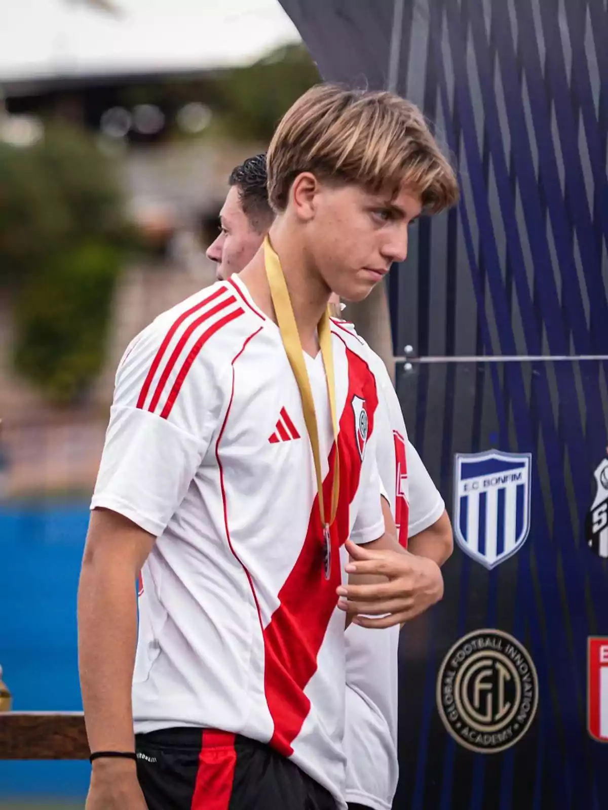 A young man with a gold medal around his neck is wearing a white jersey with a diagonal red stripe and is standing in front of a background with sports logos. A young man with a gold medal around his neck is wearing a white jersey with a diagonal red stripe and is standing in front of a background with sports logos.