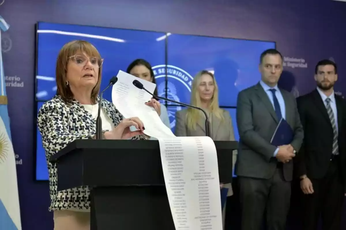 Una mujer en un podio sosteniendo un documento largo, acompañada por varias personas en un entorno formal con banderas y un logotipo oficial de fondo.