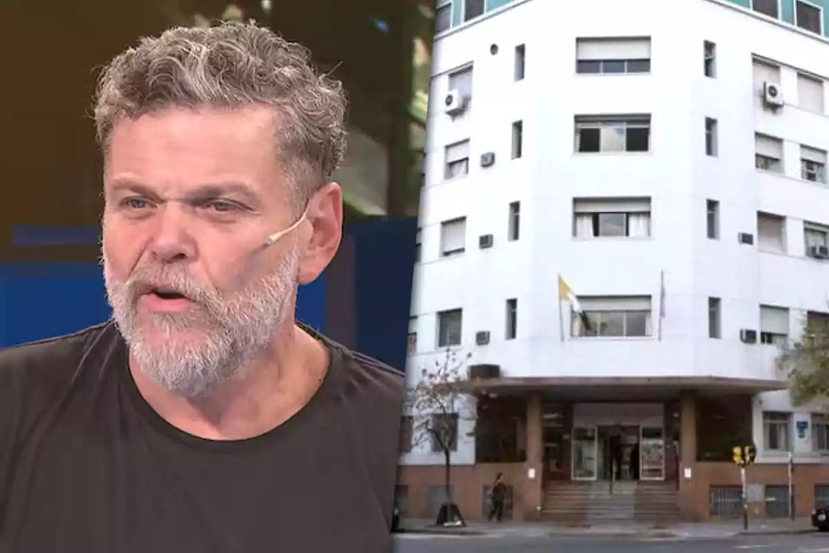 A man with a beard and a microphone in a television studio next to the image of a white multi-story building.