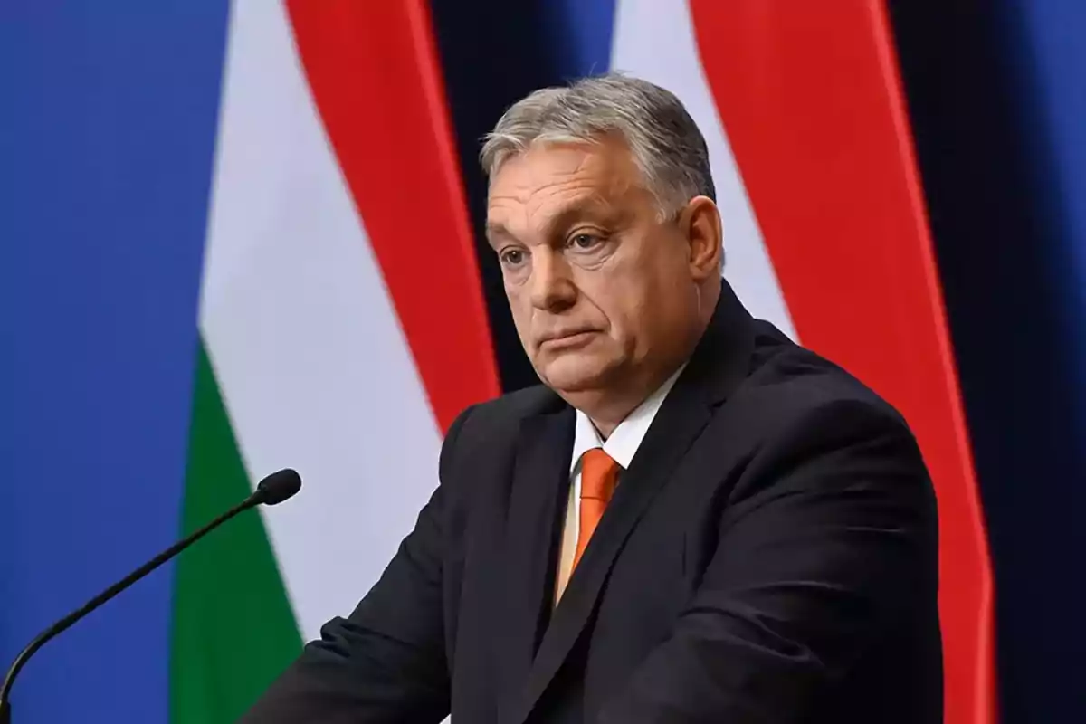 A man in a dark suit with an orange tie speaks in front of a microphone with flags in the background.