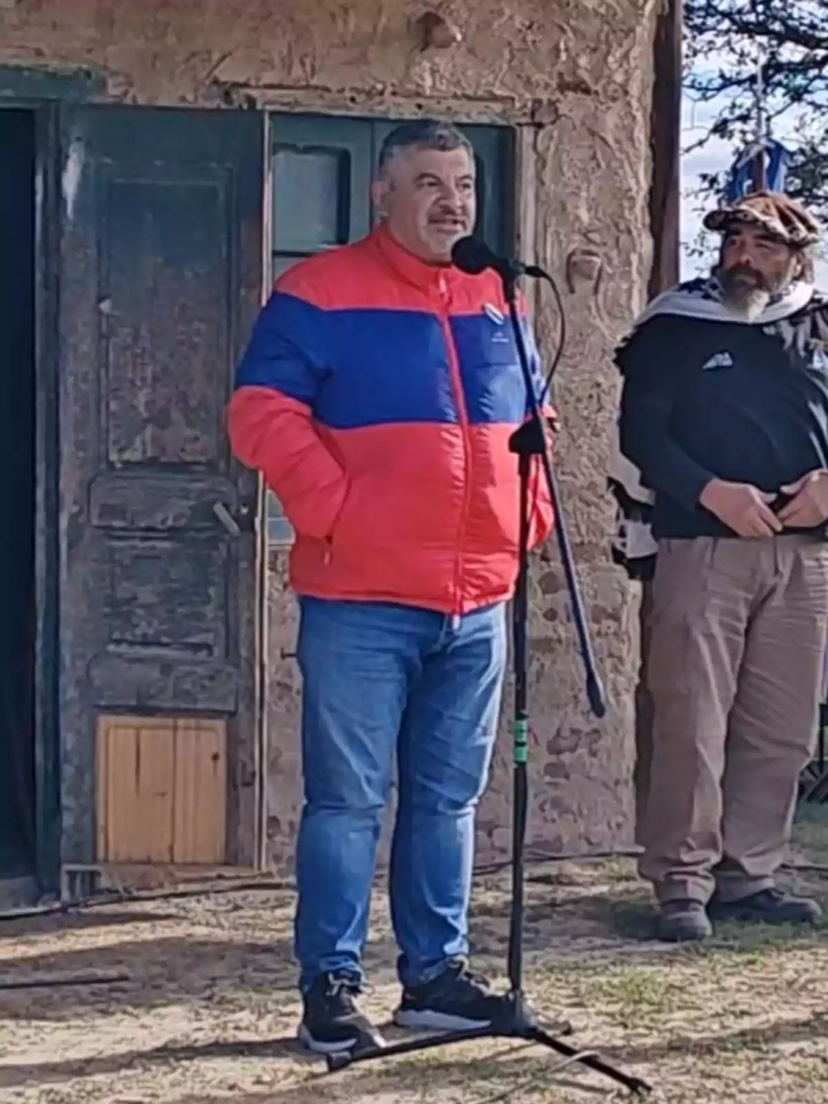 Un hombre con chaqueta roja y azul habla frente a un micrófono al aire libre, mientras otra persona está de pie a su lado. Un hombre con chaqueta roja y azul habla frente a un micrófono al aire libre, mientras otra persona está de pie a su lado.