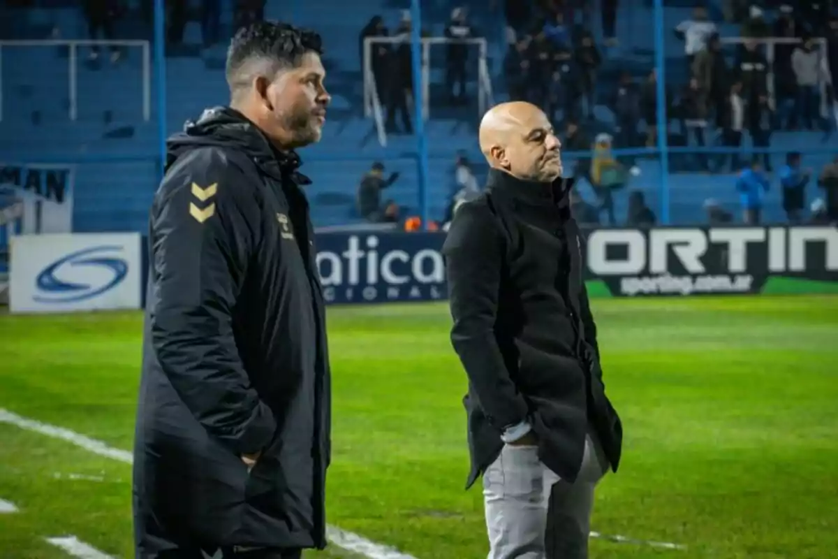 Dos entrenadores de fútbol observan el partido desde la línea de banda en un estadio con gradas azules y público al fondo Dos entrenadores de fútbol observan el partido desde la línea de banda en un estadio con gradas azules y público al fondo
