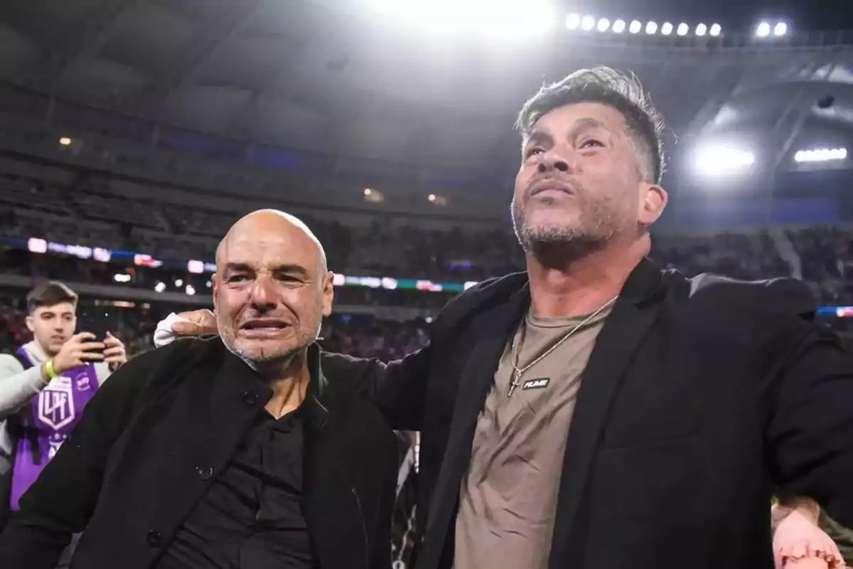 Dos hombres abrazados muestran emociones intensas en un estadio iluminado y lleno de gente, uno de ellos llora mientras el otro mira hacia arriba