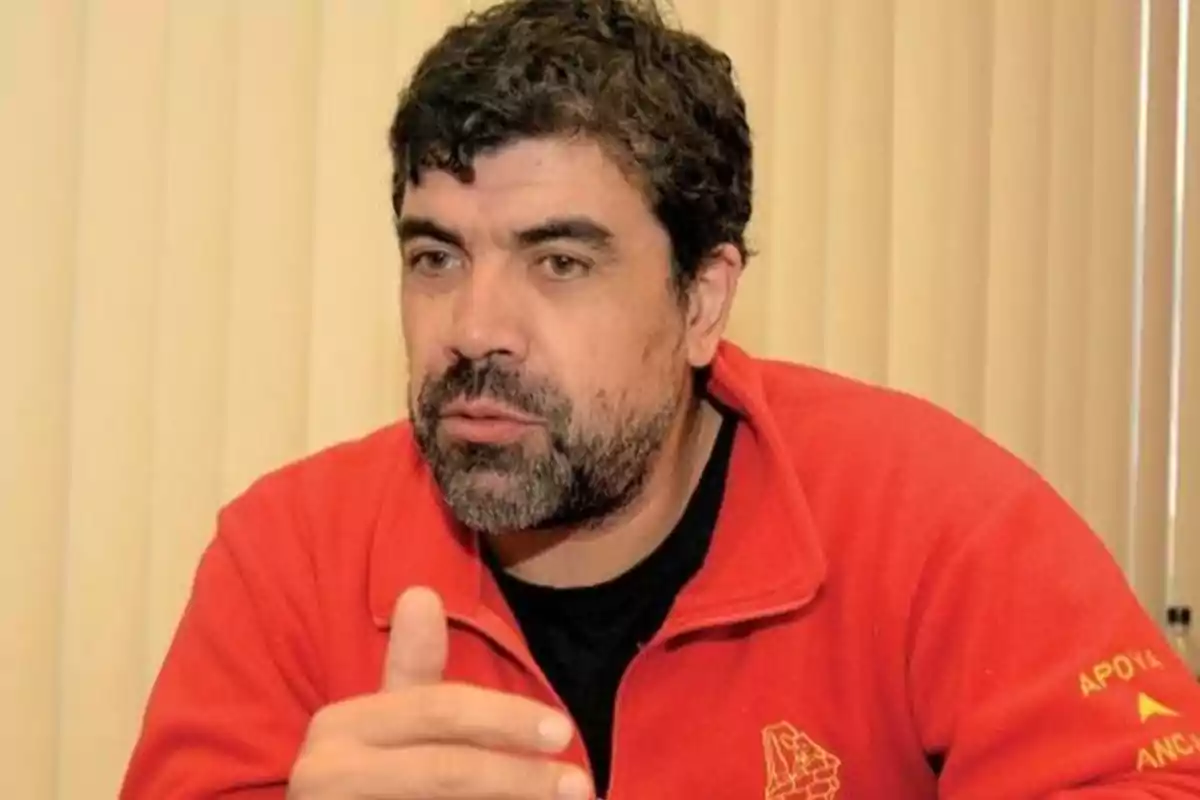 Dark-haired, bearded man in a red jacket gesturing while speaking in front of a beige blinds background Dark-haired, bearded man in a red jacket gesturing while speaking in front of a beige blinds background