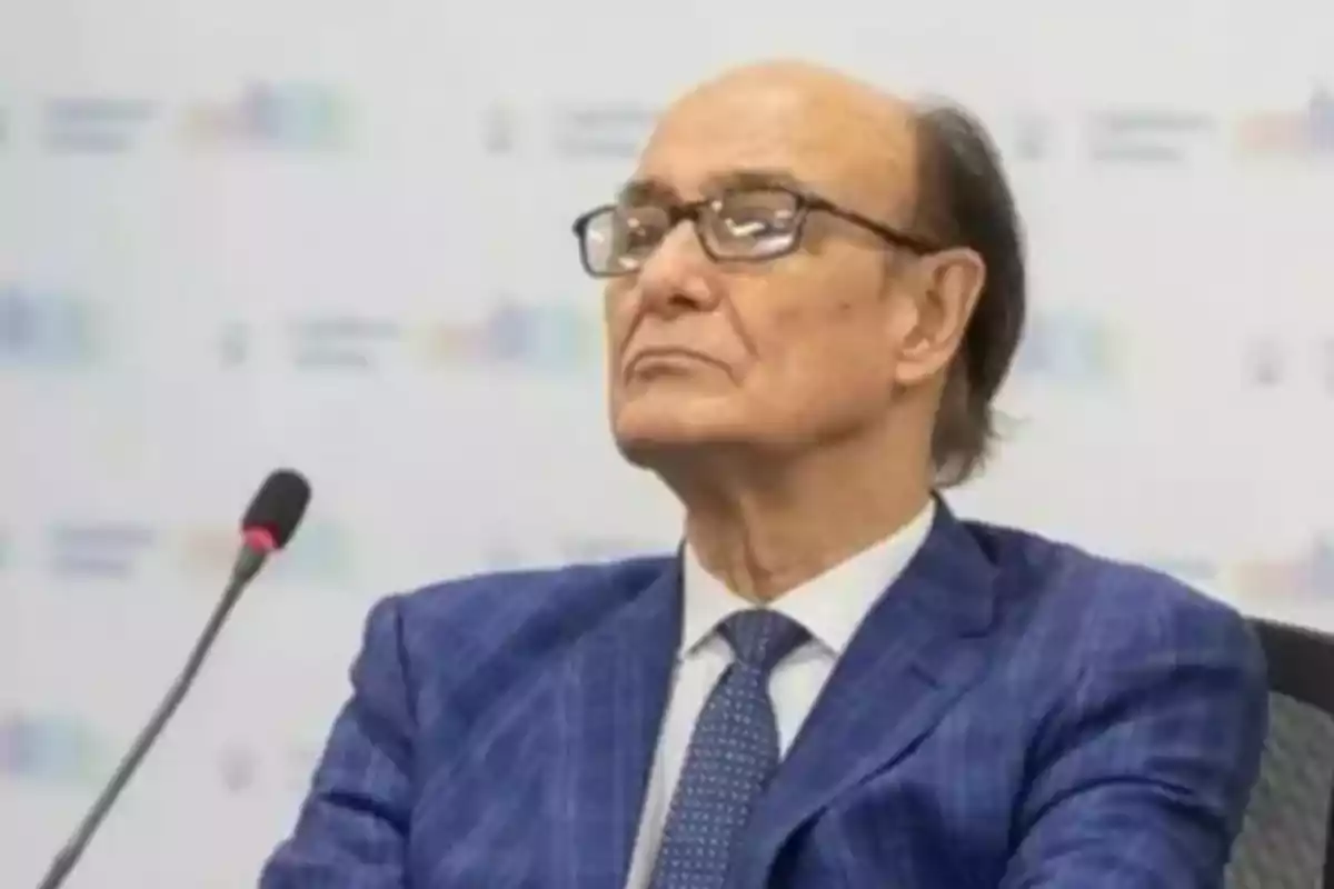 Older man with glasses and a blue suit sitting in front of a microphone in a formal setting Older man with glasses and a blue suit sitting in front of a microphone in a formal setting