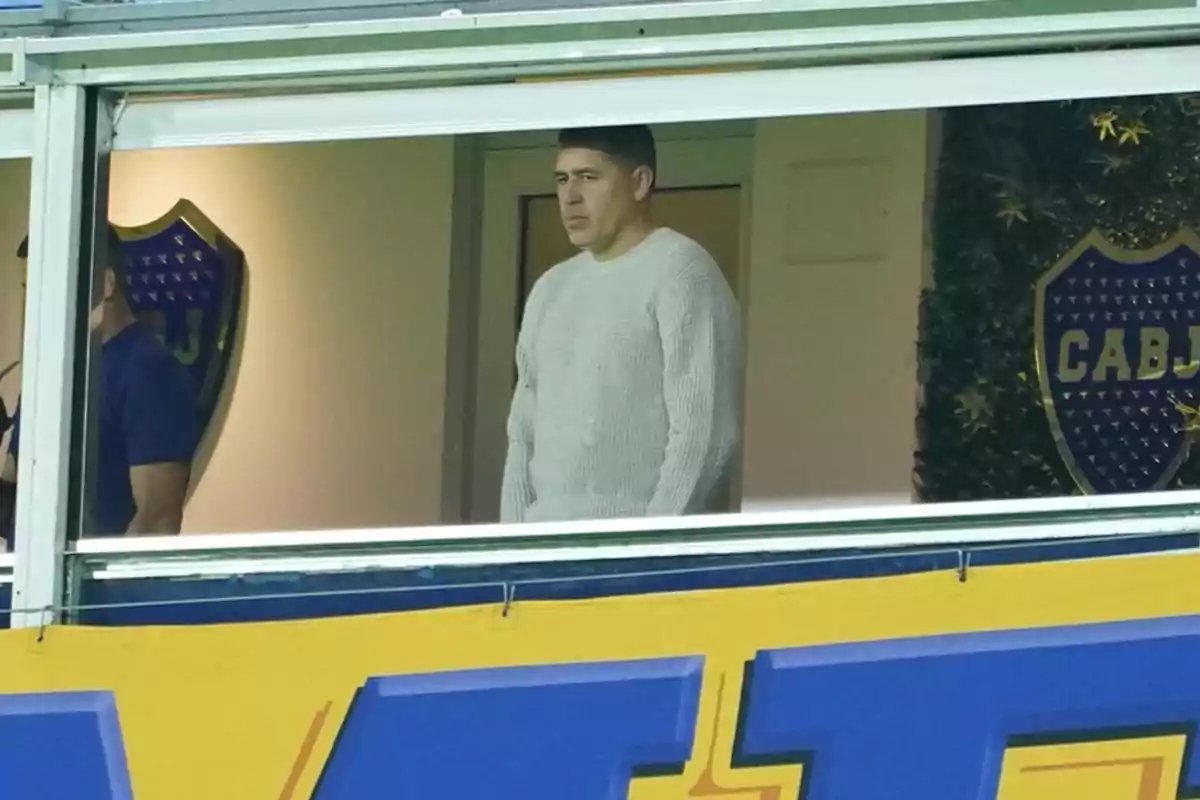 A man in a gray sweater is standing in a box at a soccer stadium with club crests on the wall. A man in a gray sweater is standing in a box at a soccer stadium with club crests on the wall.