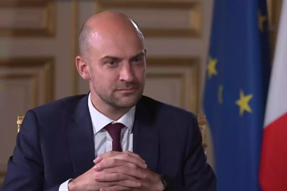 Bald man in a dark suit and burgundy tie sitting with his hands clasped in front of flags and an elegant wall Bald man in a dark suit and burgundy tie sitting with his hands clasped in front of flags and an elegant wall