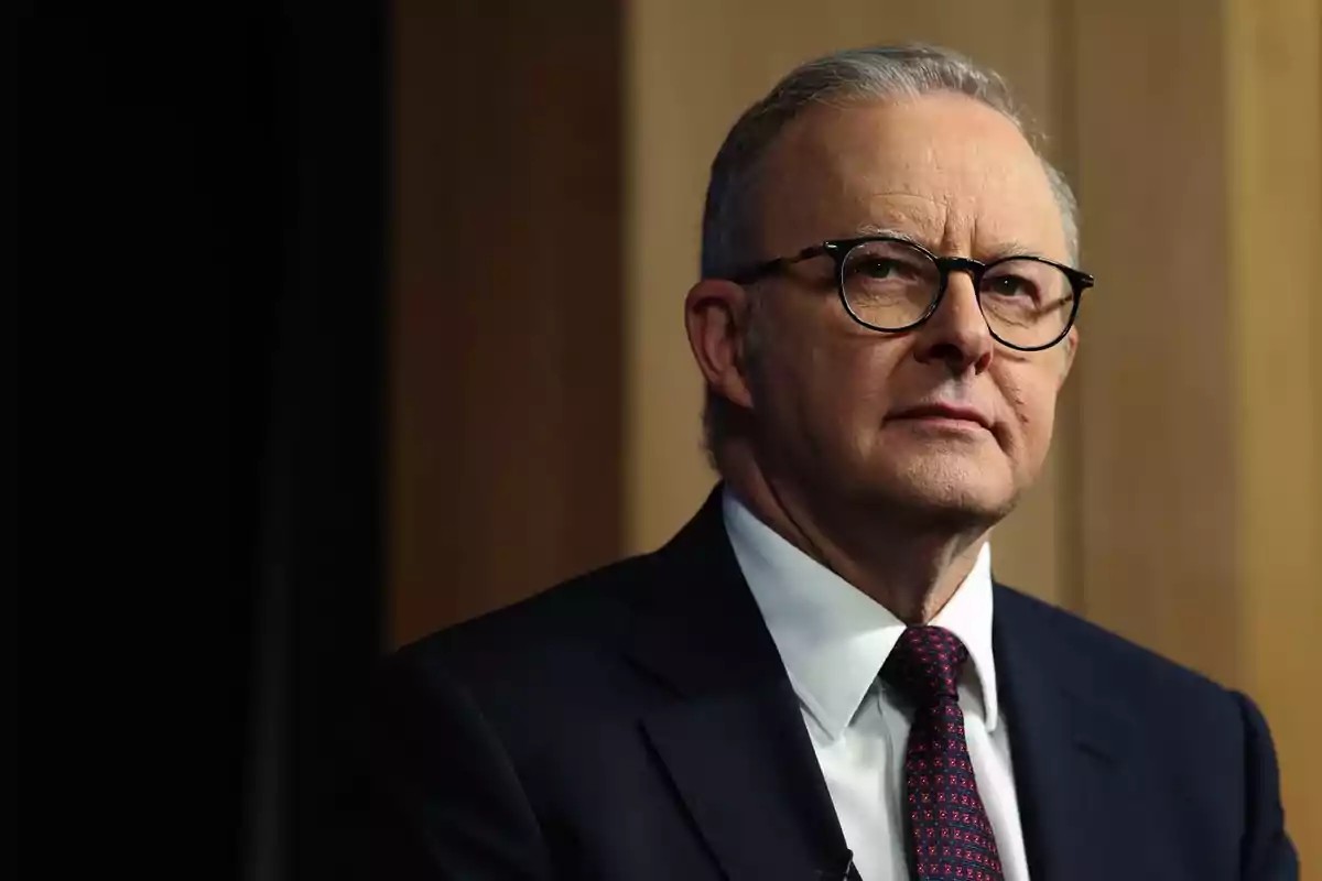 A man with glasses and a dark suit looks forward with a serious expression.