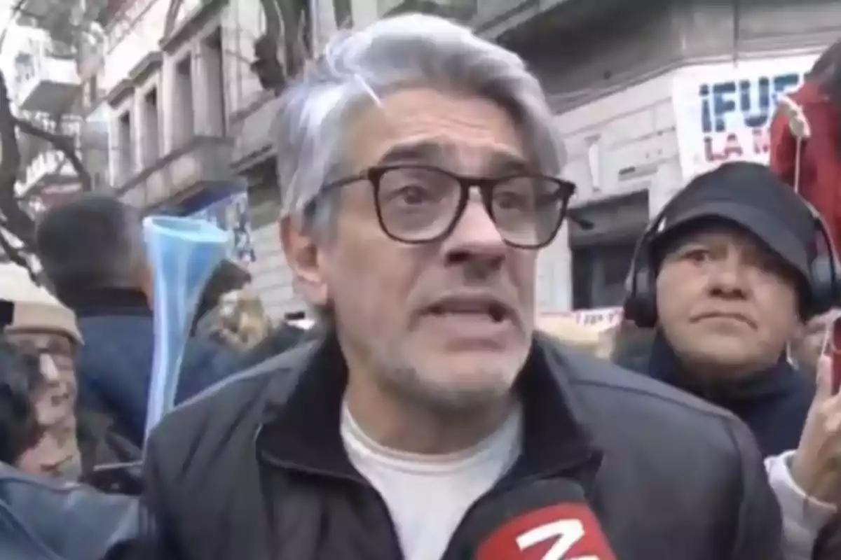 Hombre de cabello canoso y gafas hablando frente a un micrófono en una manifestación en la calle con personas y edificios al fondo