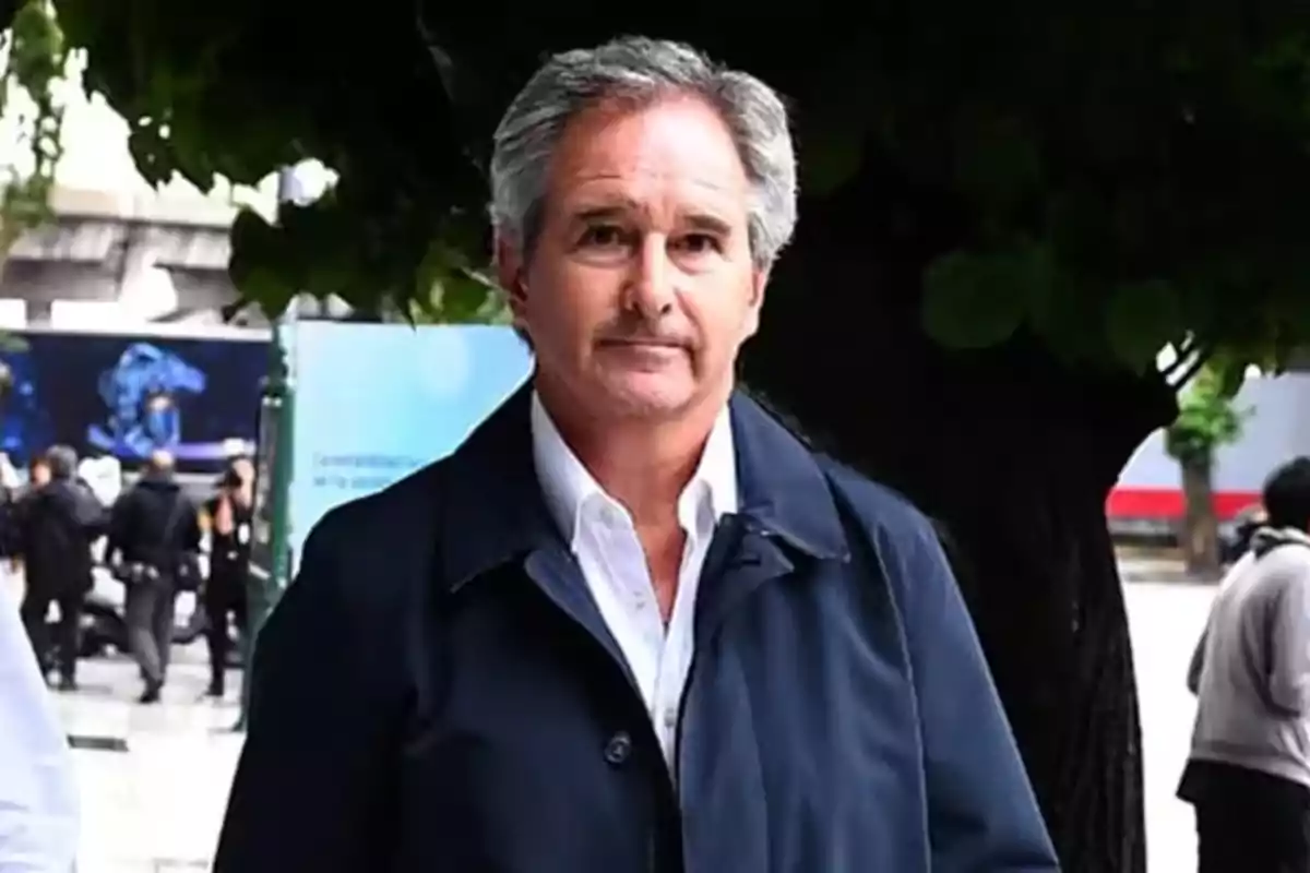 Hombre de cabello canoso con chaqueta oscura posando frente a un árbol en un entorno urbano.