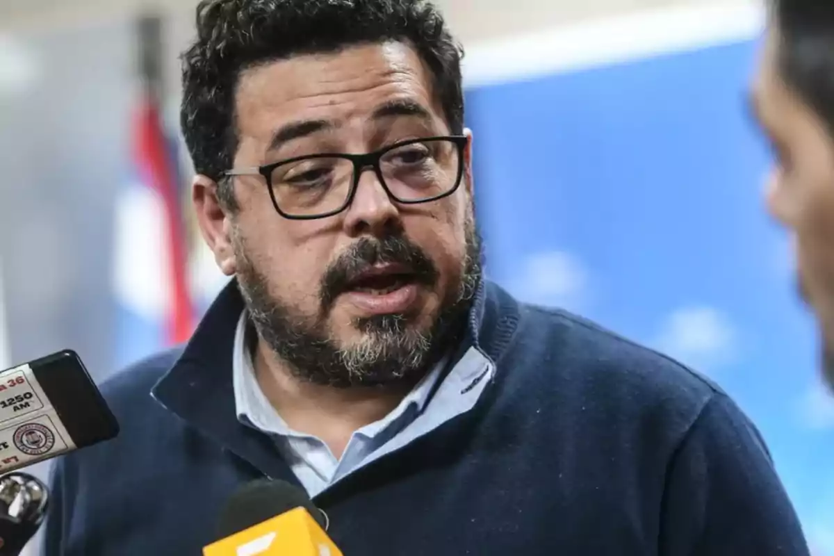 A man with glasses and a beard speaking in front of several microphones.