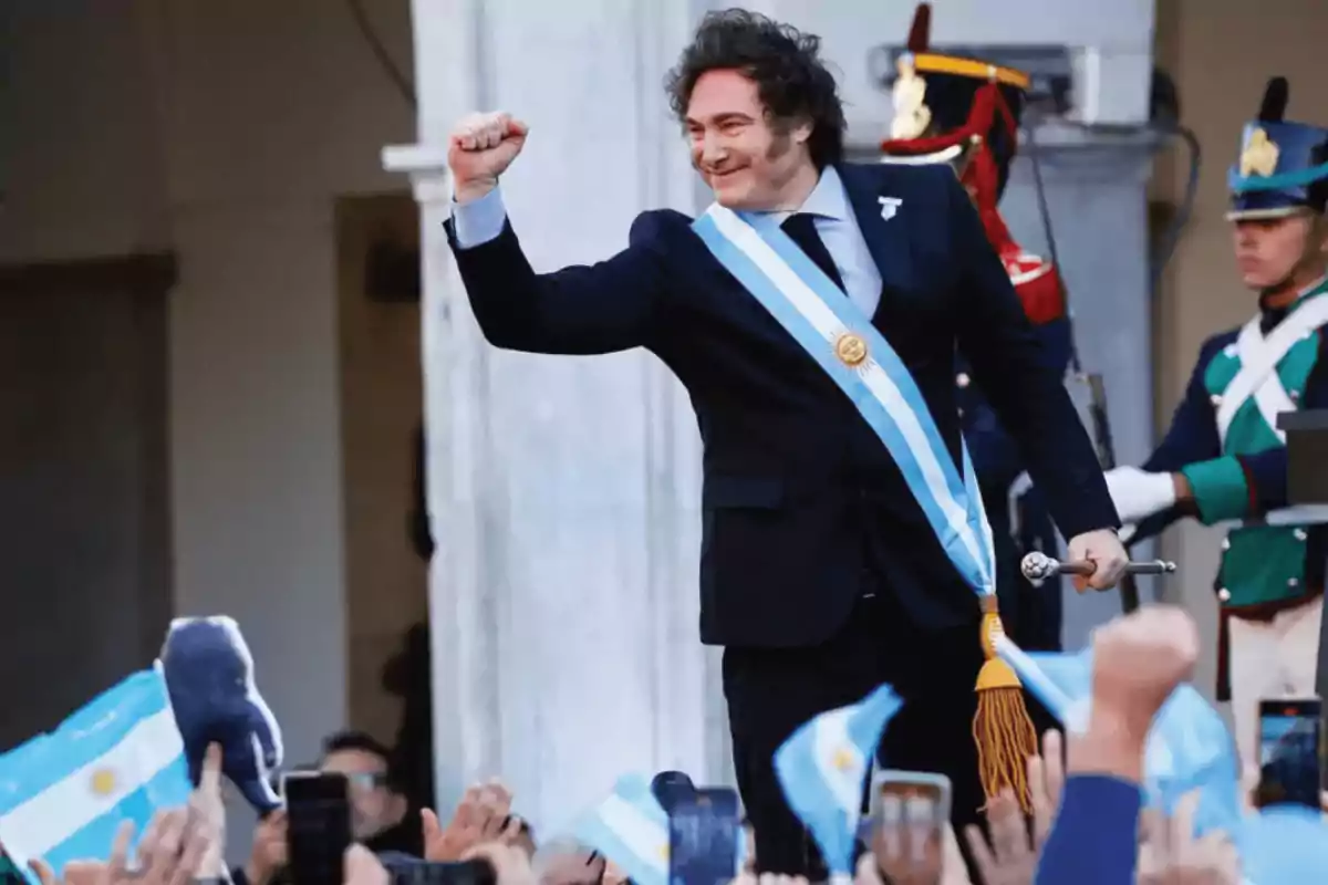 Un hombre con traje y banda presidencial levanta el puño en señal de celebración mientras una multitud ondea banderas argentinas.