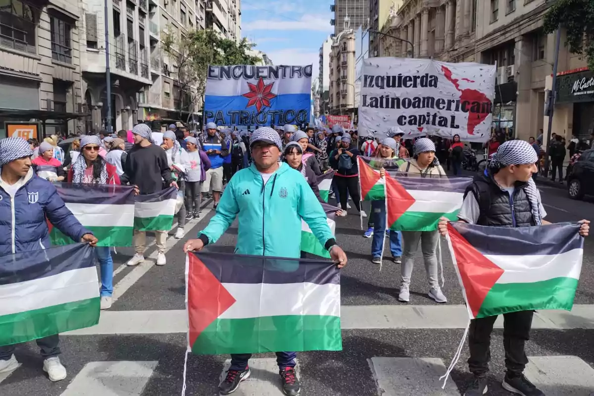 Personas marchando en una calle de la ciudad sosteniendo banderas de Palestina y usando pañuelos en la cabeza, con pancartas de fondo que tienen mensajes políticos