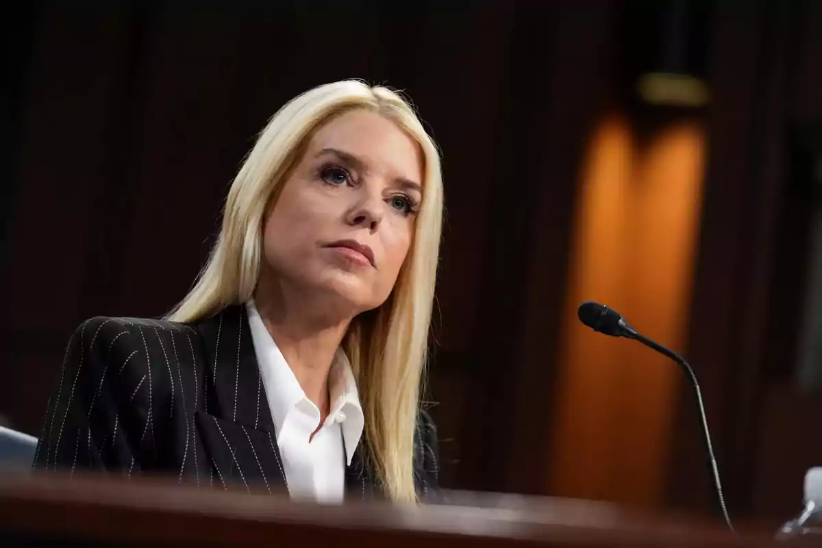 A woman with blonde hair and a dark suit is sitting in front of a microphone in a formal setting.