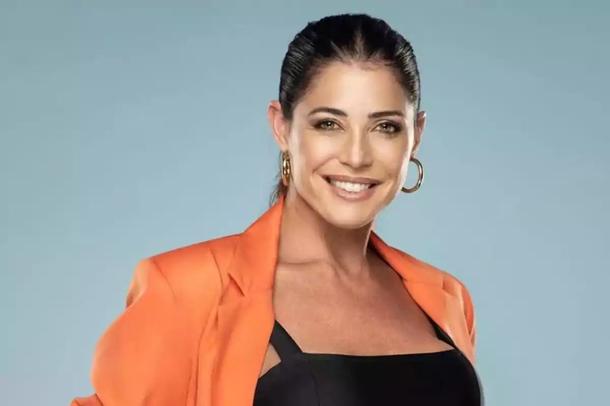 Mujer sonriendo con cabello recogido, usando un blazer naranja y aretes grandes, sobre un fondo azul claro.