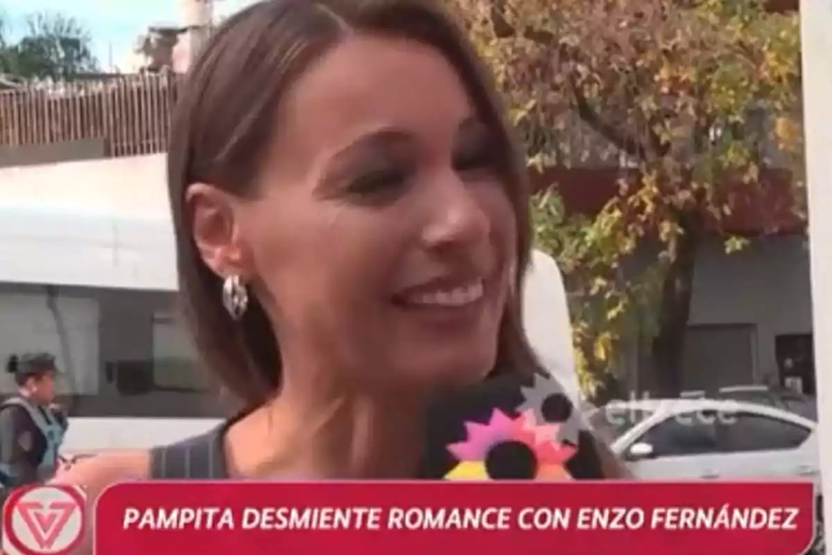 Una mujer sonriente sostiene un micrófono con un fondo urbano y un texto en la parte inferior que menciona un desmentido de romance. Una mujer sonriente sostiene un micrófono con un fondo urbano y un texto en la parte inferior que menciona un desmentido de romance.
