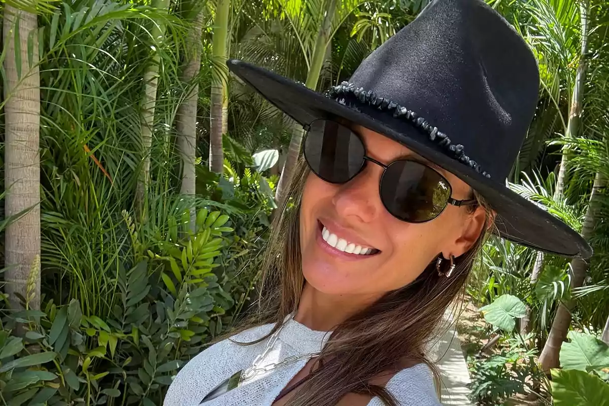 Persona con sombrero negro y gafas de sol sonriendo en un entorno tropical con palmeras y vegetación.