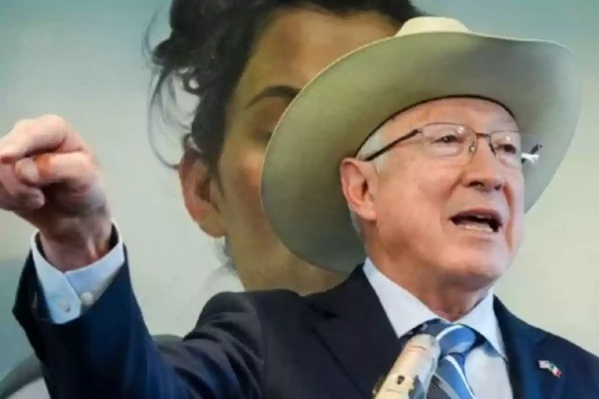 A man with a hat and glasses speaks in front of a microphone with an image in the background.