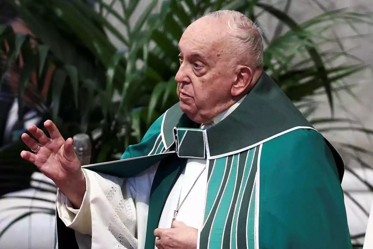 Un hombre mayor vestido con una túnica verde y blanca, levantando una mano, con plantas en el fondo.