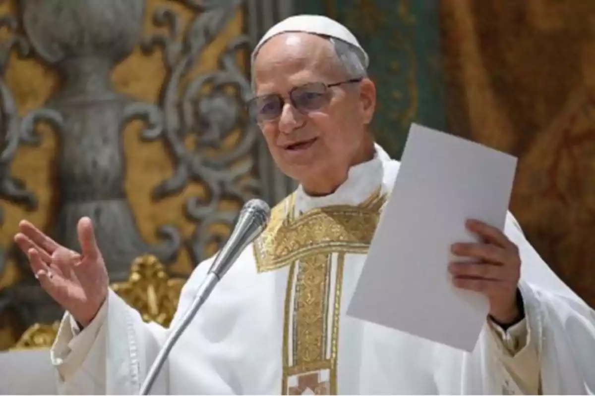 A cleric in white and gold religious attire holds a paper while speaking in front of a microphone in a decorated setting. A cleric in white and gold religious attire holds a paper while speaking in front of a microphone in a decorated setting.