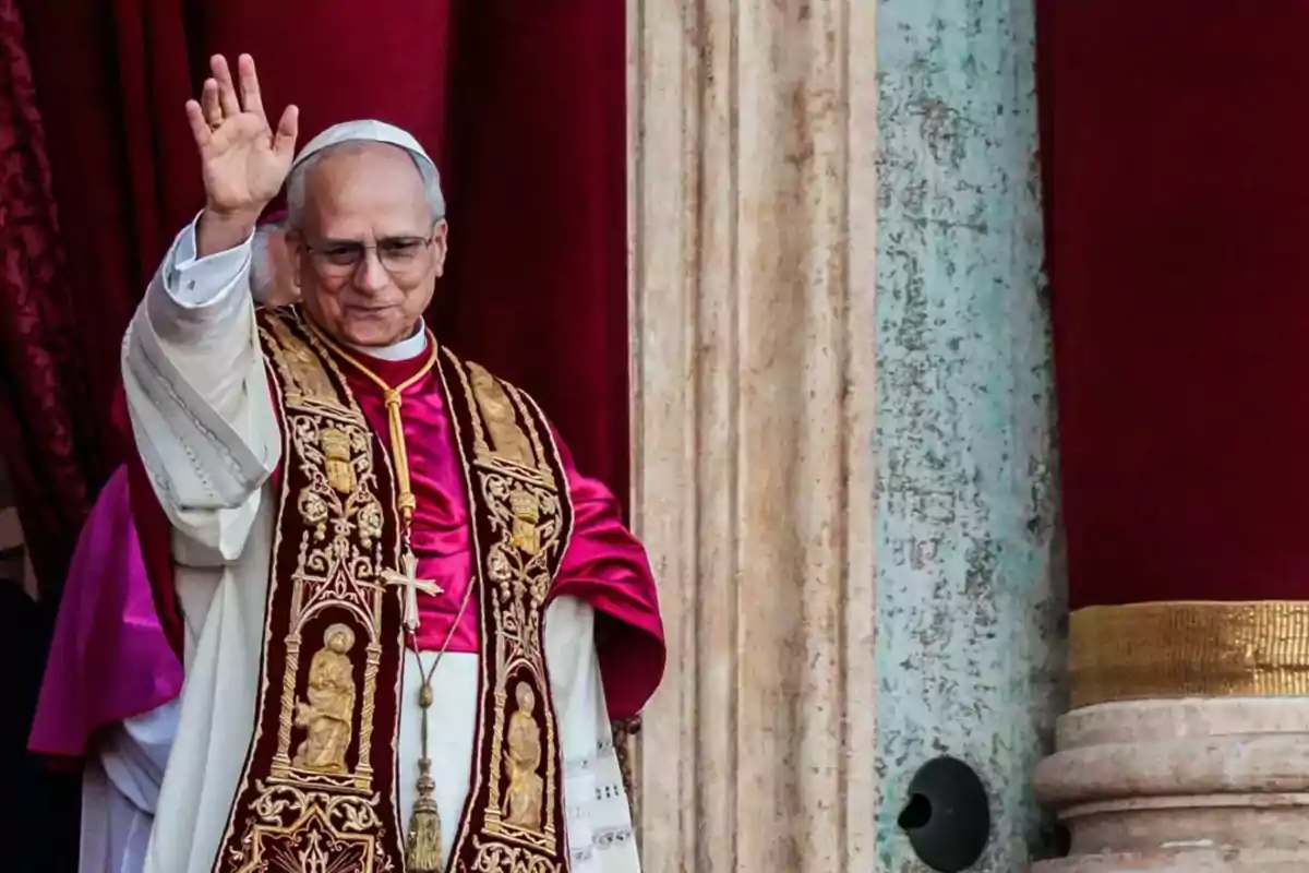 A man dressed in elaborate religious attire, including a white cassock and an embroidered cape, raises his hand in a gesture of greeting while standing in front of a backdrop of red curtains. A man dressed in elaborate religious attire, including a white cassock and an embroidered cape, raises his hand in a gesture of greeting while standing in front of a backdrop of red curtains.