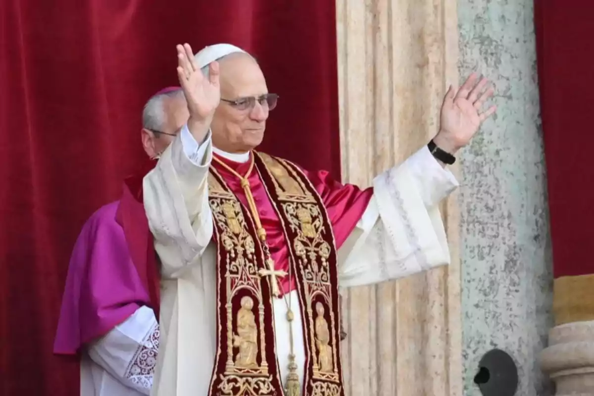 Un hombre vestido con vestimenta religiosa levanta las manos en señal de saludo frente a un fondo rojo. Un hombre vestido con vestimenta religiosa levanta las manos en señal de saludo frente a un fondo rojo.