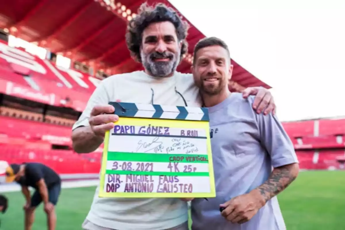 Two people smile and pose in a soccer stadium while one of them holds a clapperboard. Two people smile and pose in a soccer stadium while one of them holds a clapperboard.