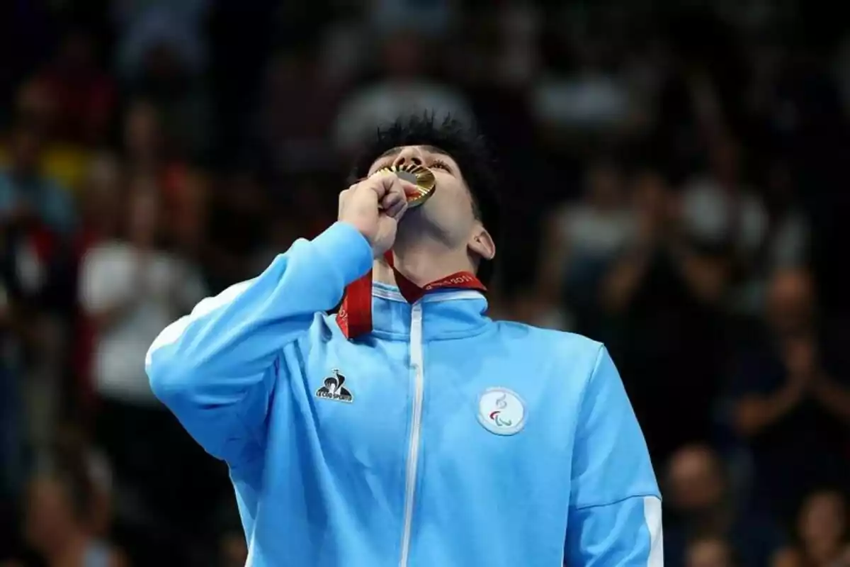 Un atleta con una chaqueta azul claro besa una medalla de oro.