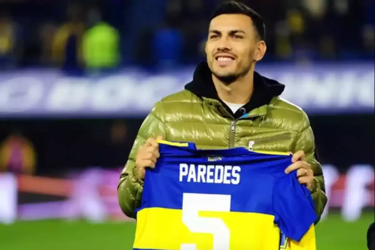 Un futbolista sonriente sostiene una camiseta azul y amarilla con el nombre Paredes y el número 5 en un estadio de fútbol. Un futbolista sonriente sostiene una camiseta azul y amarilla con el nombre Paredes y el número 5 en un estadio de fútbol.
