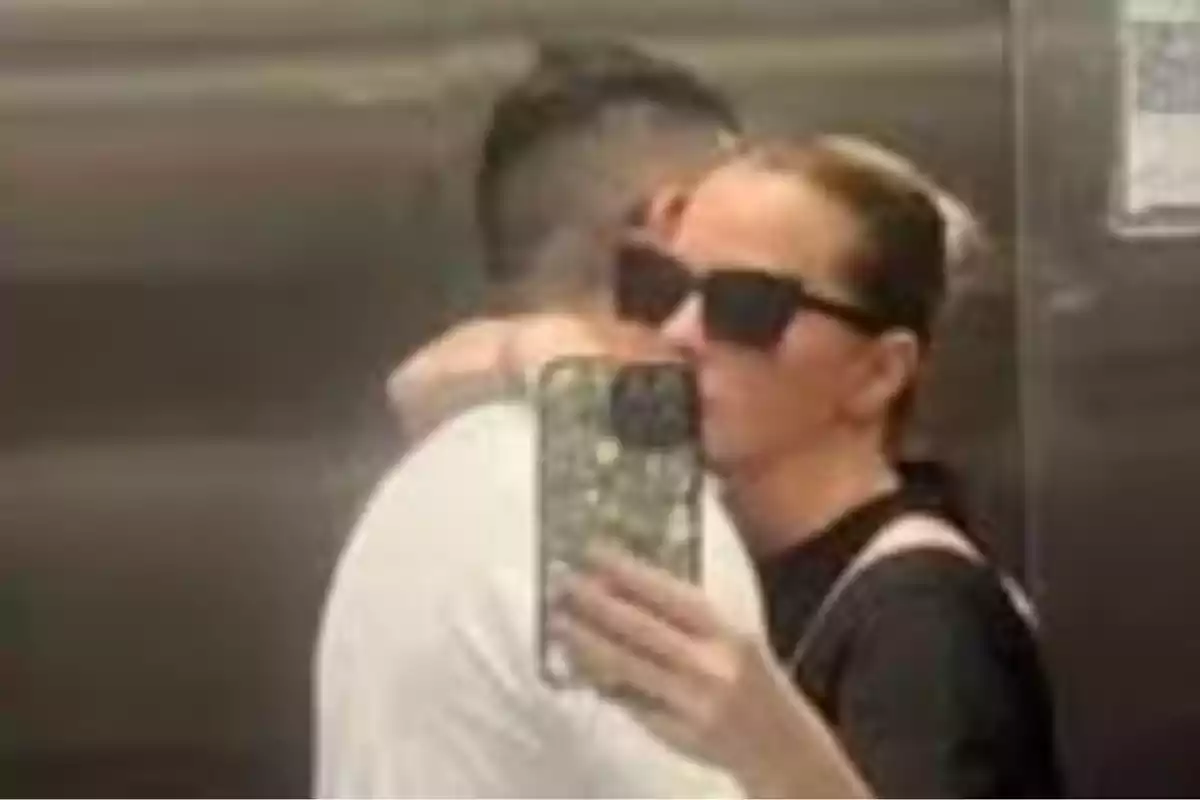 A person with sunglasses takes a selfie in a mirror while hugging another person in an elevator. A person with sunglasses takes a selfie in a mirror while hugging another person in an elevator.