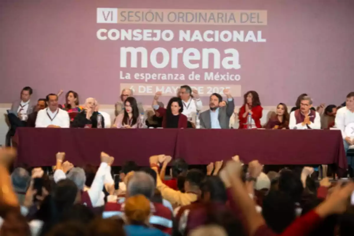 Un grupo de personas está sentado en una mesa larga frente a una audiencia que levanta los puños en señal de apoyo, con un fondo que muestra un cartel de un evento político. Un grupo de personas está sentado en una mesa larga frente a una audiencia que levanta los puños en señal de apoyo, con un fondo que muestra un cartel de un evento político.