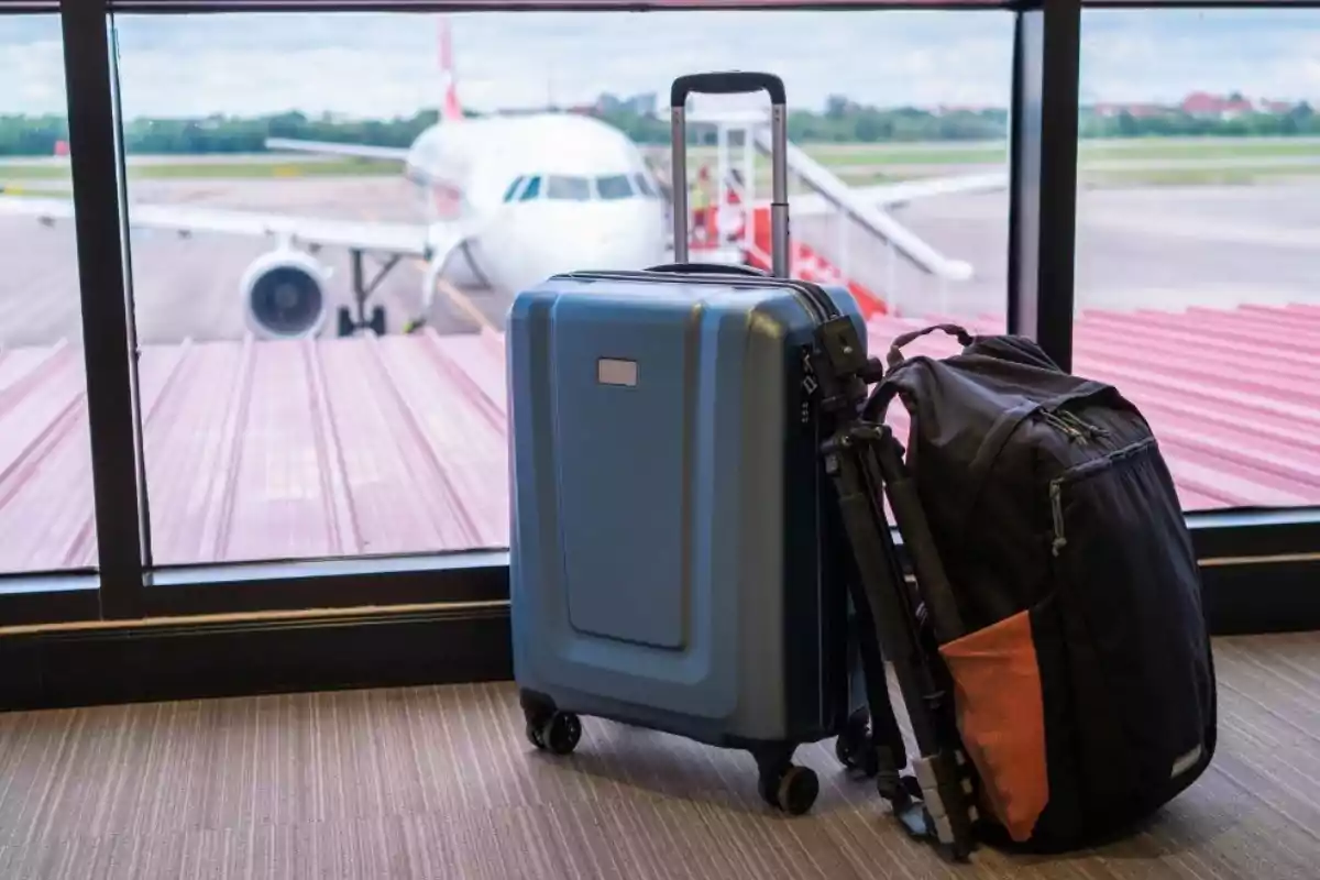 Una maleta azul y una mochila negra están junto a una ventana en un aeropuerto con un avión en la pista al fondo. Una maleta azul y una mochila negra están junto a una ventana en un aeropuerto con un avión en la pista al fondo.