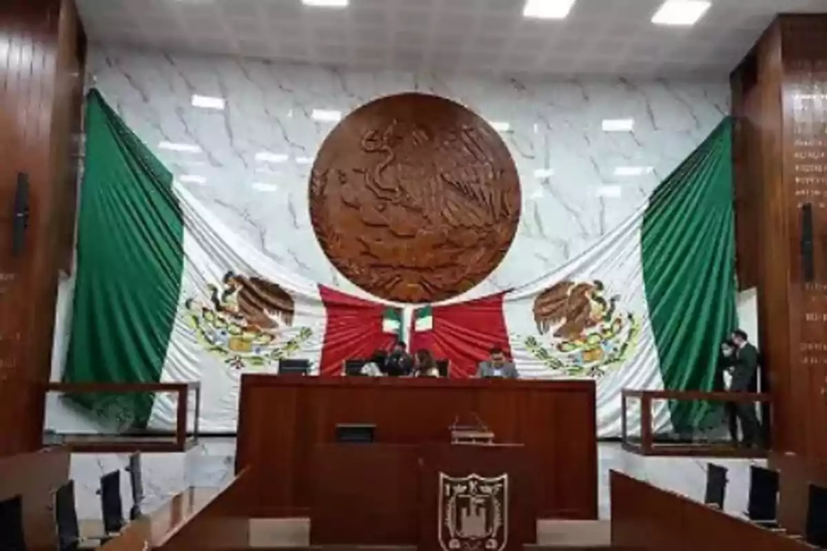 Salón de sesiones con una gran bandera de México y un escudo en la pared detrás de un podio.