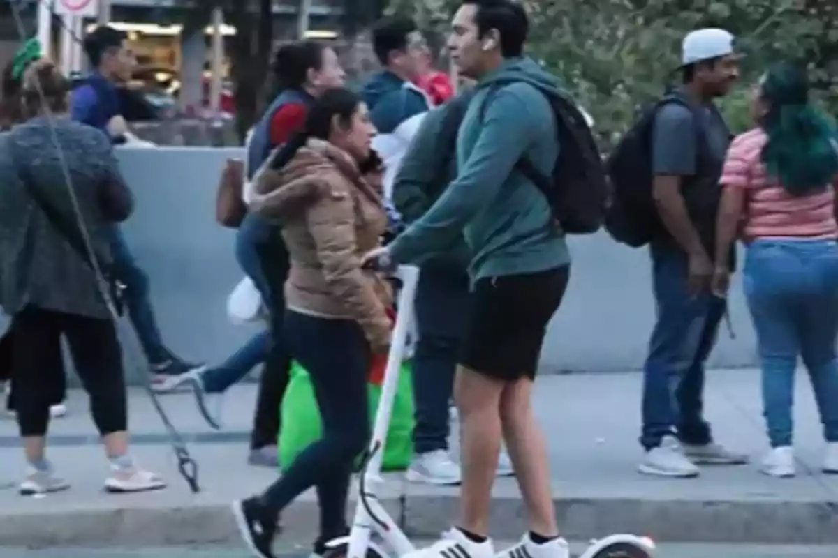 Un hombre en patineta eléctrica y una mujer caminando se cruzan en una acera concurrida.