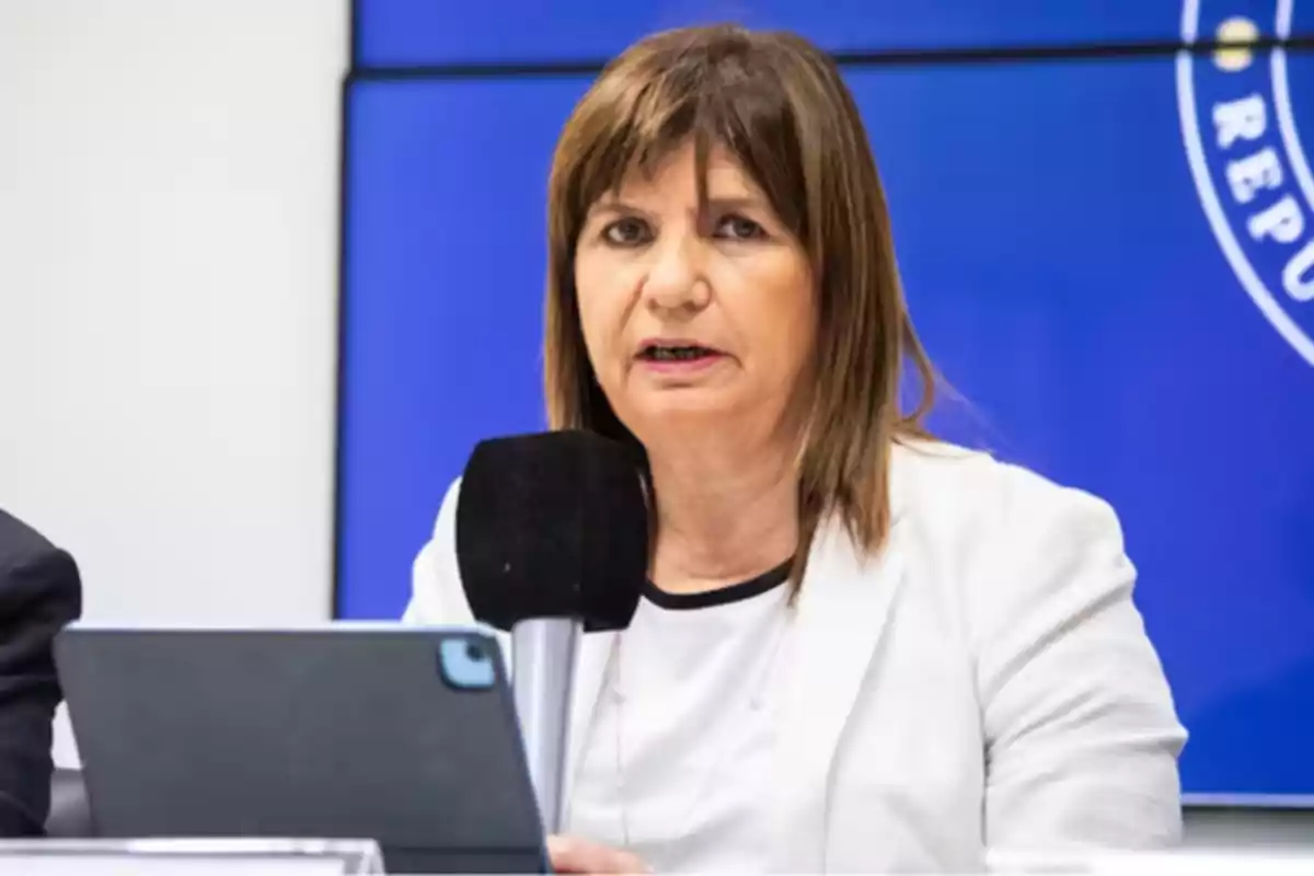 A person speaking at a press conference with a microphone and a tablet in front of a blue background.