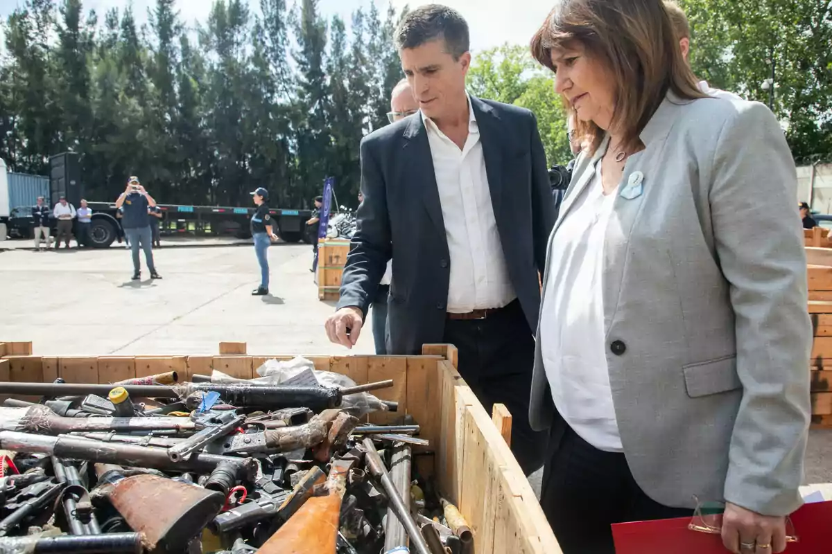 Dos personas observan un contenedor lleno de armas decomisadas en un espacio al aire libre con otras personas y vehículos al fondo Dos personas observan un contenedor lleno de armas decomisadas en un espacio al aire libre con otras personas y vehículos al fondo