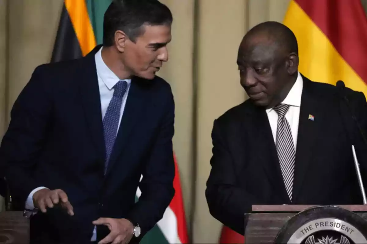 Dos hombres de traje conversando en un evento oficial con banderas de fondo.