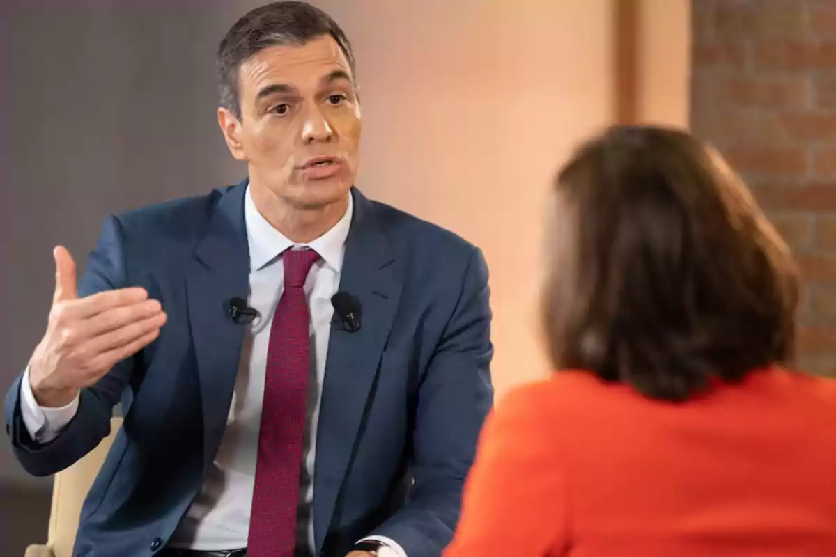 Un hombre en traje azul y corbata roja hablando con una mujer de espaldas en una entrevista.