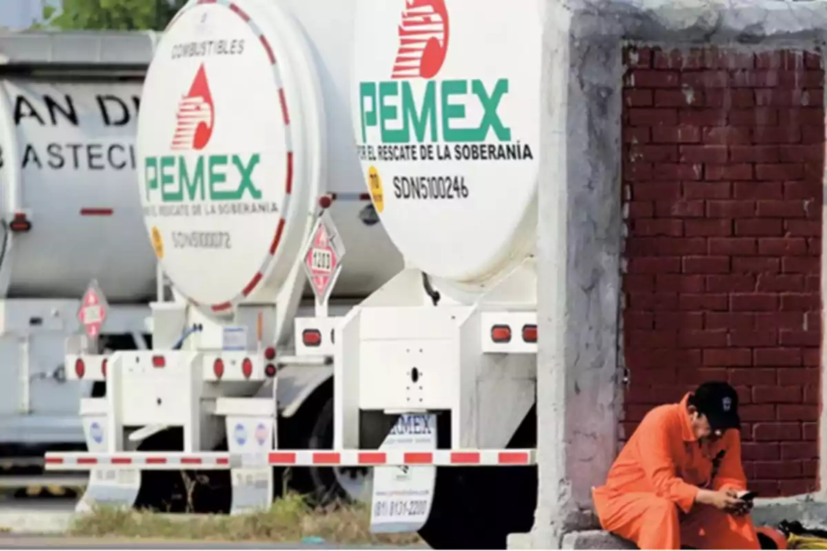 Un trabajador con uniforme naranja revisa su teléfono mientras está sentado junto a un muro de ladrillos cerca de camiones cisterna de PEMEX estacionados.