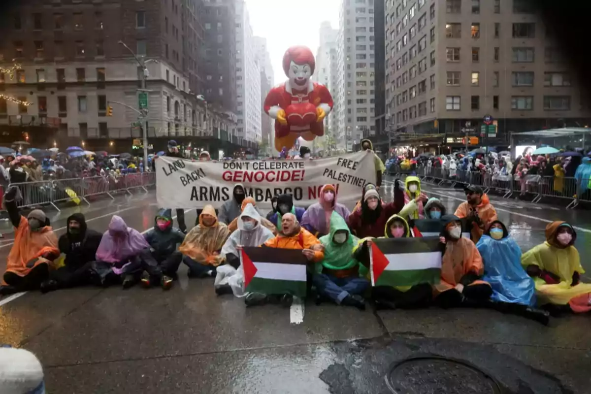 Un grupo de personas con impermeables de colores sostiene pancartas y banderas en una calle lluviosa durante una manifestación, mientras un globo gigante de un personaje de comida rápida flota en el fondo.