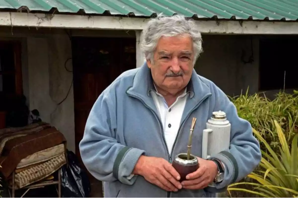 Un hombre mayor con cabello canoso sostiene un mate y un termo frente a una casa con techo de chapa verde. Un hombre mayor con cabello canoso sostiene un mate y un termo frente a una casa con techo de chapa verde.