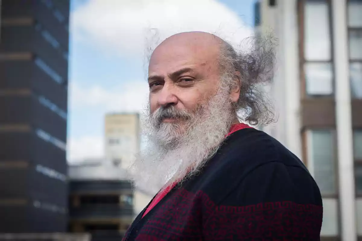 A man with a white beard and messy hair looks at the camera with an urban background of buildings.