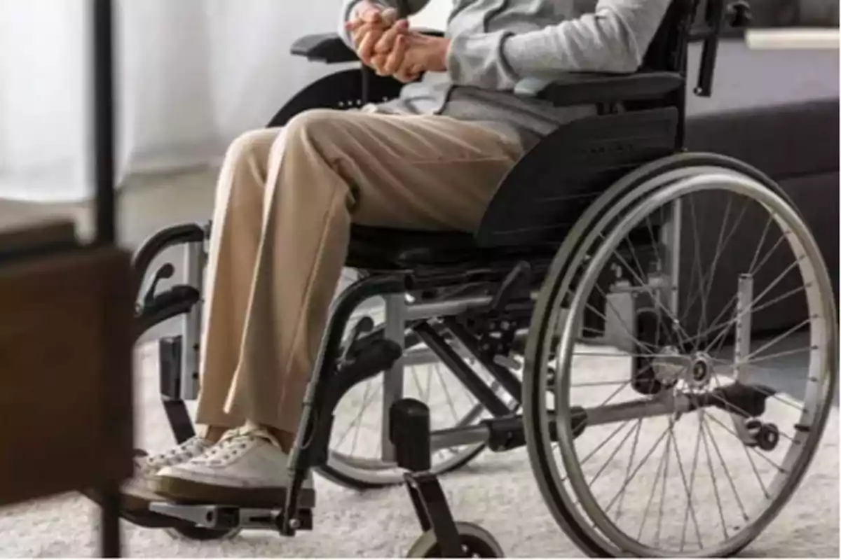 Person sitting in a wheelchair with hands clasped, wearing beige pants and white shoes.