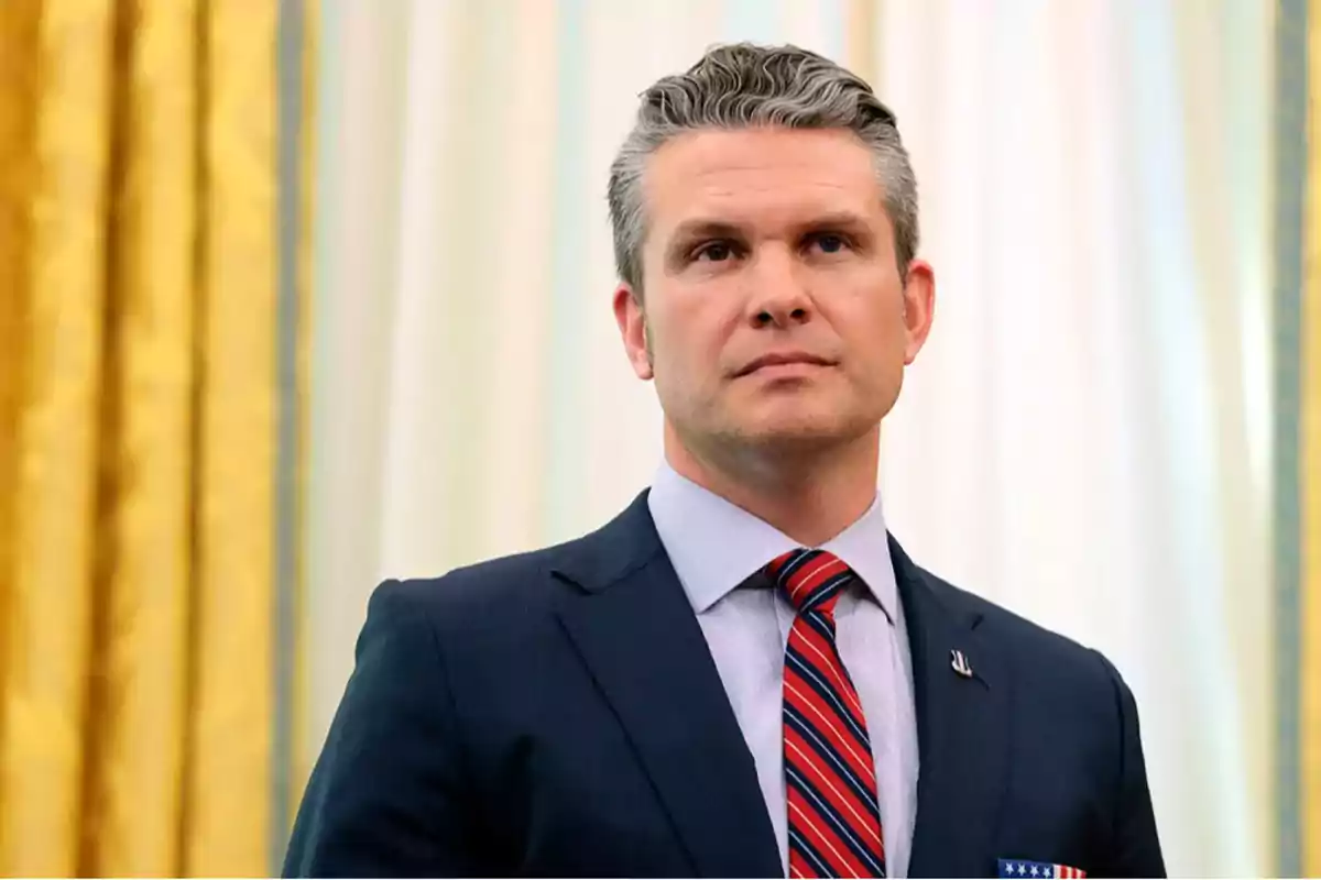 Un hombre de cabello canoso y traje oscuro con corbata a rayas rojas y azules está de pie frente a un fondo con cortinas doradas y blancas. Un hombre de cabello canoso y traje oscuro con corbata a rayas rojas y azules está de pie frente a un fondo con cortinas doradas y blancas.