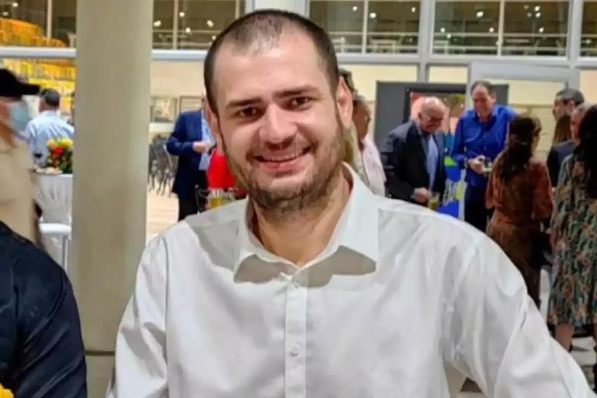 A smiling man in a white jersey at a social event with people in the background. A smiling man in a white jersey at a social event with people in the background.
