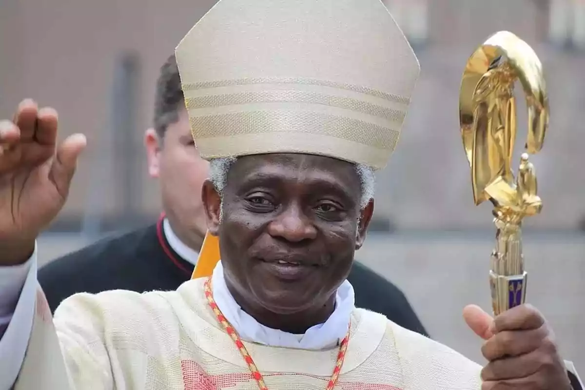 A Catholic cleric in ceremonial attire with a golden staff smiles while raising one hand. A Catholic cleric in ceremonial attire with a golden staff smiles while raising one hand.